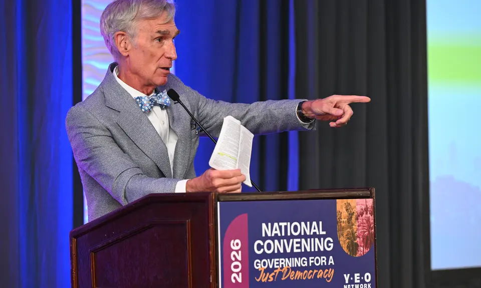 Bill Nye wearing a gray suit and blue bow tie standing at a podium at the Young Elected Officials Network Convening 2026 and holding a copy of the Constitution. 