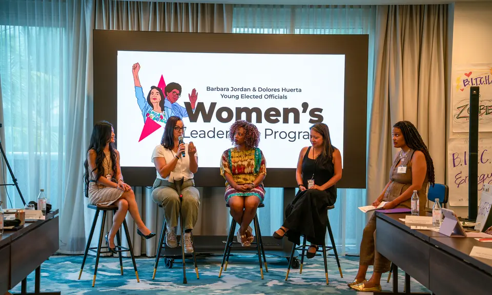 Five women sit on stools in front of a sign that reads "Women's Leadership Program"
