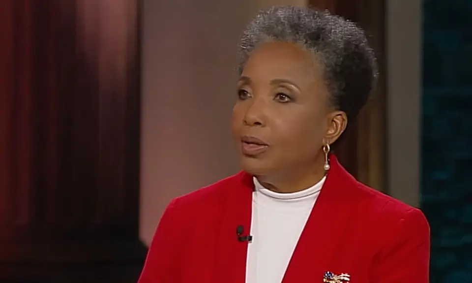 Carol Swain, a Black woman with graying hair and wearing a white shirt under a bright red jacket, is seated on set and looking toward her off-camera interviewer