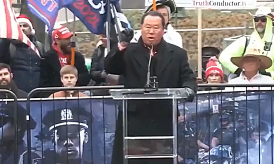 Che Ahn speaking at microphone at outdoors Jan. 5, 2021 rally on Freedom Plaza near the White House. Ahn is wearing black coat and gloves, people holding Trump flags are visible behind him.