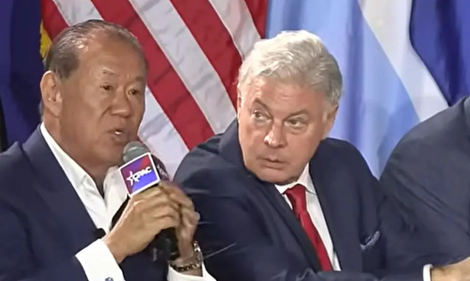 Che Ahn speaks into a microphone branded with a CPAC logo; seated to his left and listening to Ahn is Lance Wallnau.