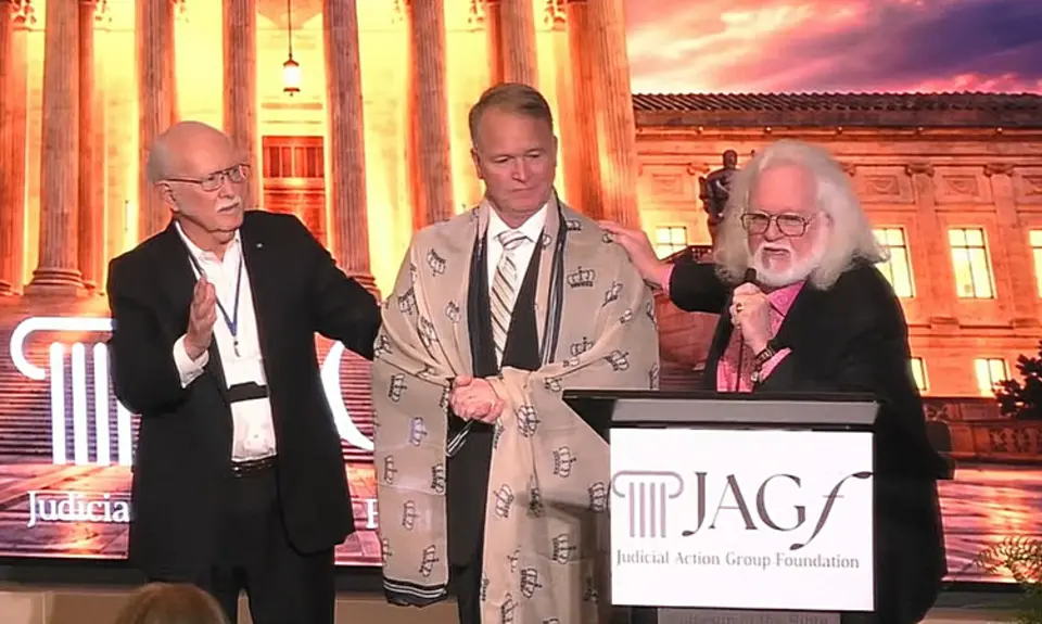 Dominionist Chuck Pierce, who has a gray beard and long gray hair, stands behind a lectern with the logo for the Judicial Action Group Foundation logo. His right hand rests on the shoulder of Phillip Jauregui, who has a shawl embroidered with crowns draped over his shoulders. A third man has his hand up in prayer. They are standing before an image of the U.S. Supreme Court building lit up at night. 