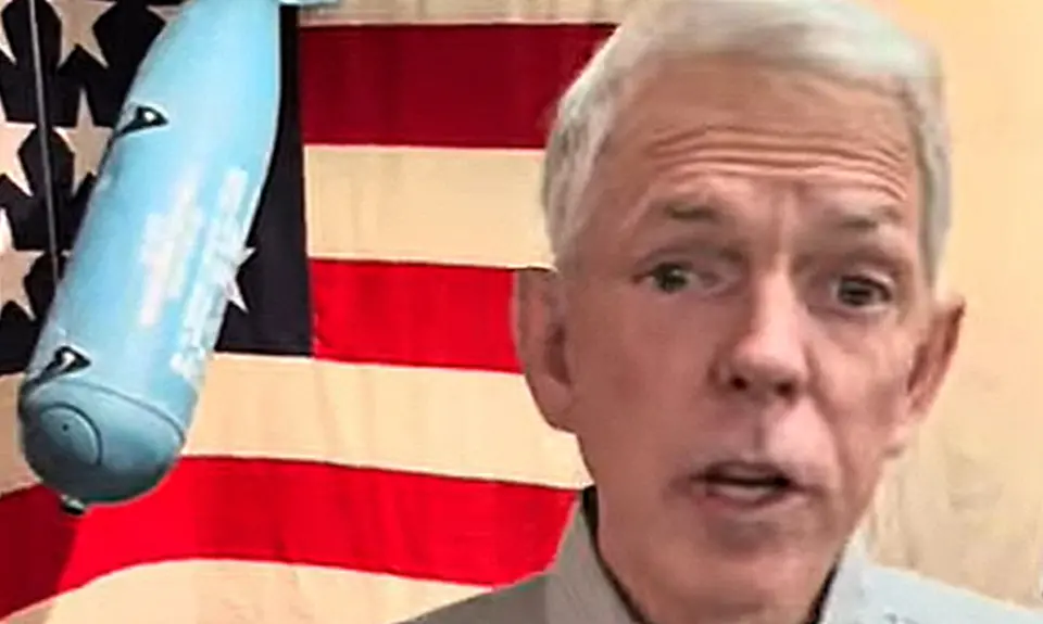 David Barton speaks into the camera; visible behind him is a World War II era (?) bomb hanging in front of a U.S. flag.