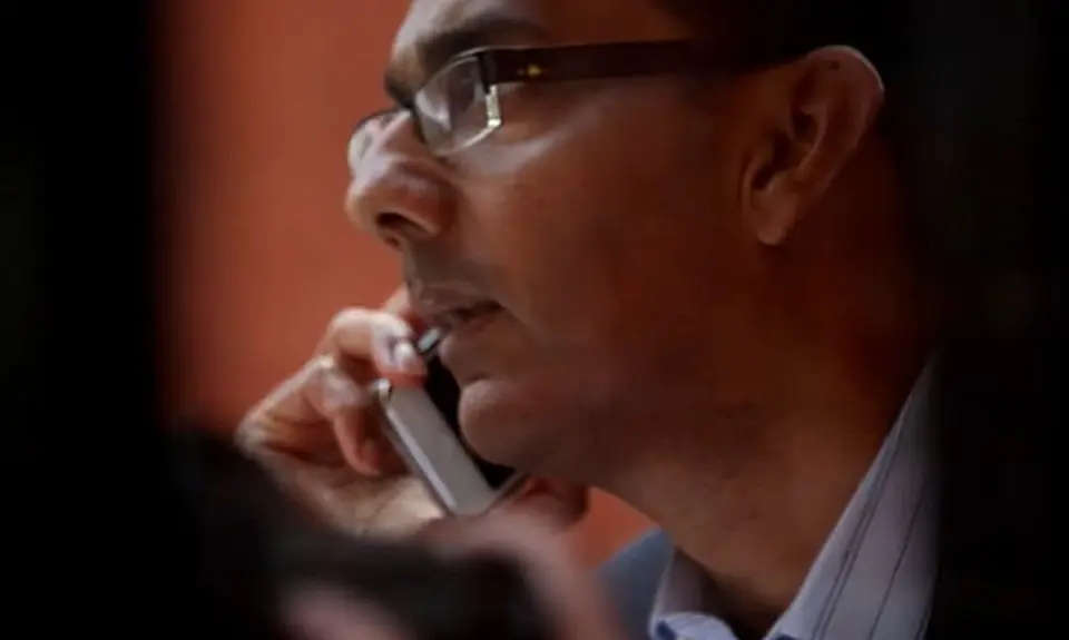 Close up side-angle view of Dinesh D'Souza looking serious and looking off camera to the left while holding a cell phone to his right ear.