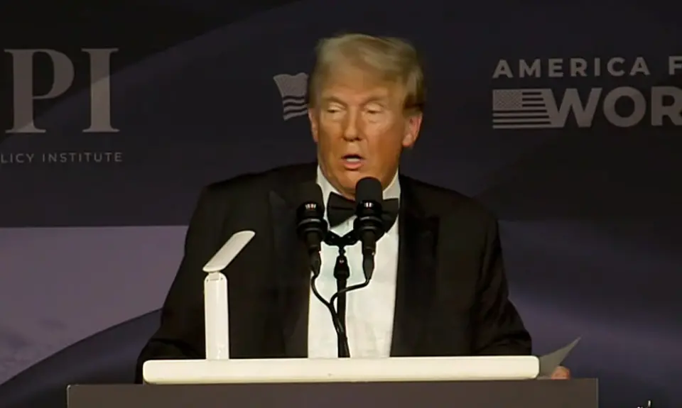 Donald Trump in a tuxedo speaks at a lectern in front of logos for the America First Policy Institute and its political arm, America First Works.
