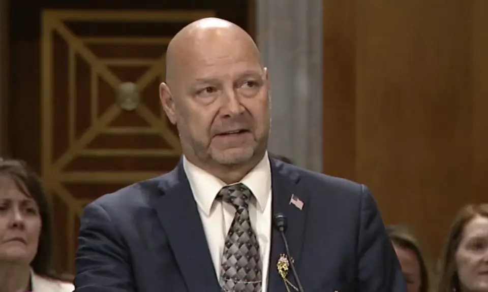 Doug Mastriano in suit and tie is seated at a table in a Senate hearing room, speaking to an off-camera senator