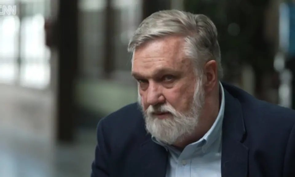 Douglas Wilson, a white man with gray hair and beard, is looking off camera to the CNN reporter interviewing him. The background is blurred. 