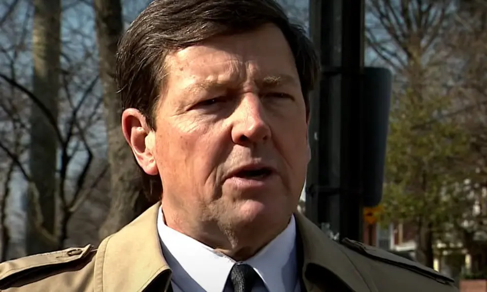 Ed Martin, wearing suit and tie and tan overcoat, is being interviewed outdoors; visible behind him are neighborhood homes and trees. 