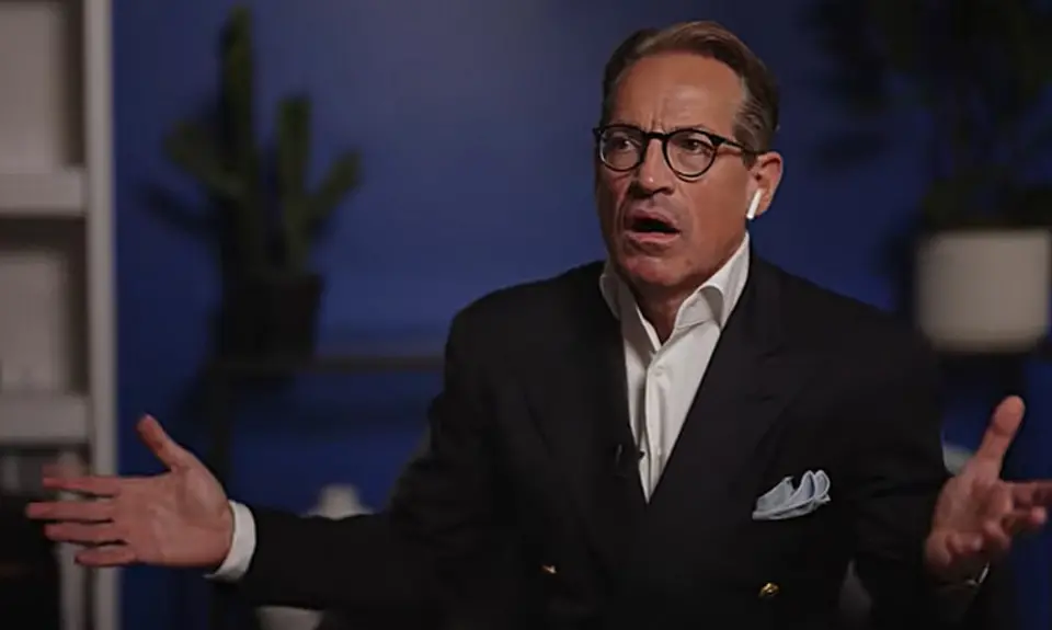 Metaxas gestures with both arms open while sitting in what appears to be a living room or home office