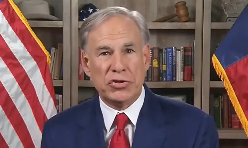 Gov. Greg Abbott wearing blue suit and red tie speaks to the camera; bookshelves are visible behind him with an American flag partially visible to the left and Texas flag to the right