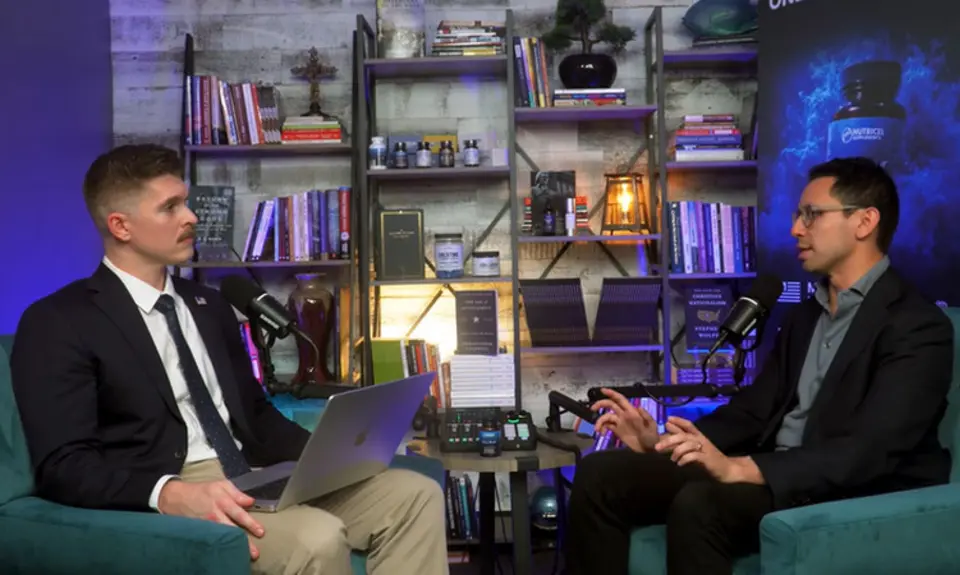 Joshua Haymes and James Fishback are seated in arm chairs facing each other in front of a set of bookshelves. Haymes has a laptop open on his lap.