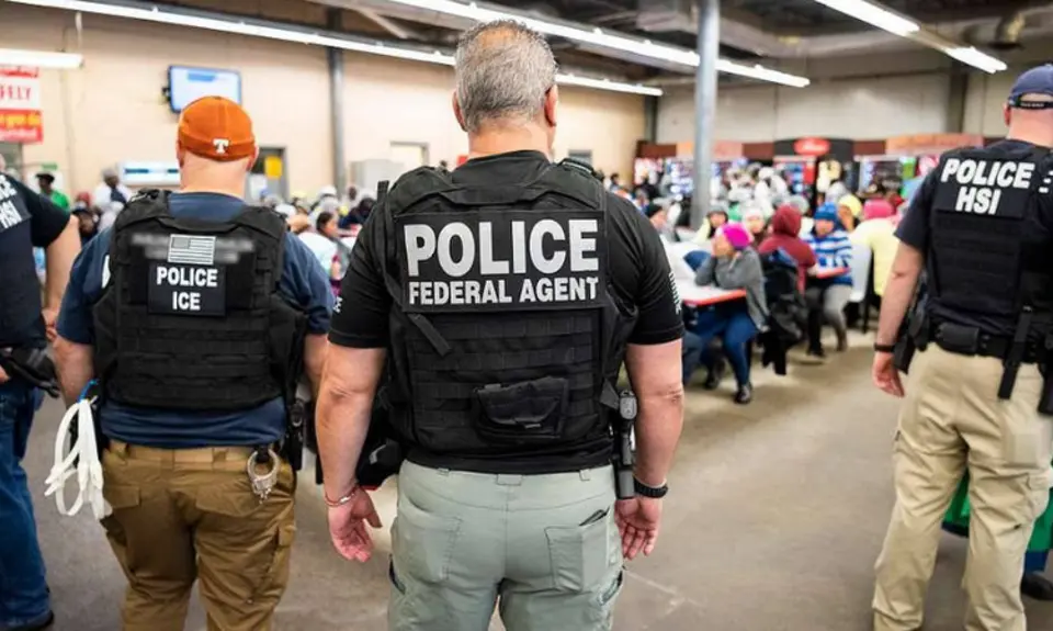 Photo shows several officers identified as police and/or ICE from the back as they face workers gathered in what appears to be a workplace lunchroom