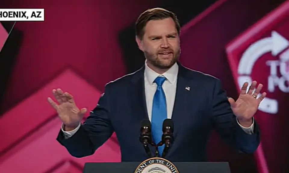 Vice President J.D. Vance gestures with his hands while speaking at lectern on stage at TPUSA's AmericaFest; the TPUSA logo is partially visible behind him.