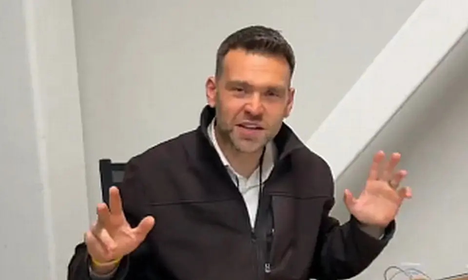 Jack Posobiec, a casually dressed white man seated at his laptop, gestures with his hands while speaking into the camera in front of a white wall background.