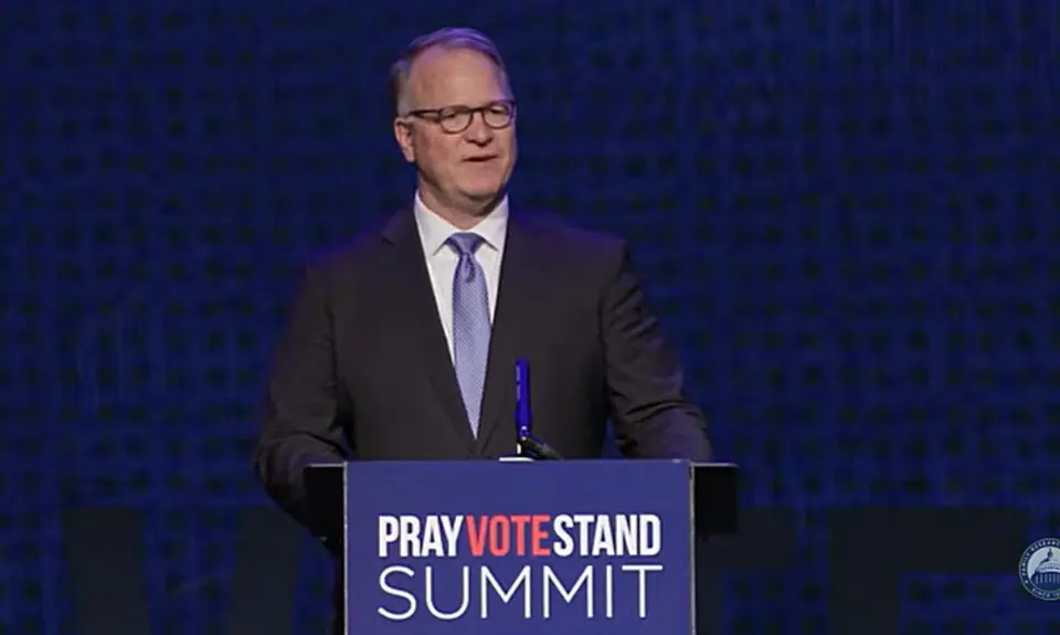 Phillip Jauregui, balding white man wearing a suit and tie, stands behind a lectern that carries the logo for the Pray Vote Stand Summit