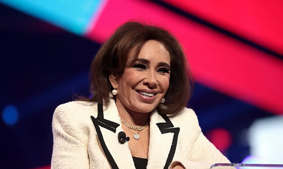 Jeanine Pirro, wearing cream colored suit with black trim, smiles while standing at lectern with red and blue stage backdrop blurred behind her.
