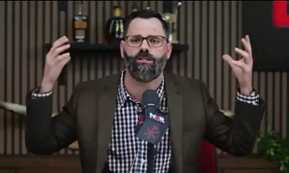 Joel Webbon gestures with both hands while seated behind a microphone that displays the logo for his NXR (New Christian Right) Studios