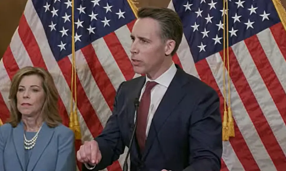 Image from press conference shows Sen. Josh Hawley in dark suit and red tie and standing in front of two U.S. flags, looking off camera at a questioner. Standing behind him on the left is Kristen Waggoner, head of the anti-abortion legal group Alliance Defending Freedom. 
