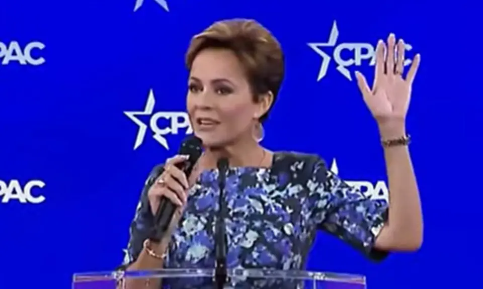 Kari Lake on stage in front of CPAC logos; she holds microphone in her right hand with her left hand raised in the air. 
