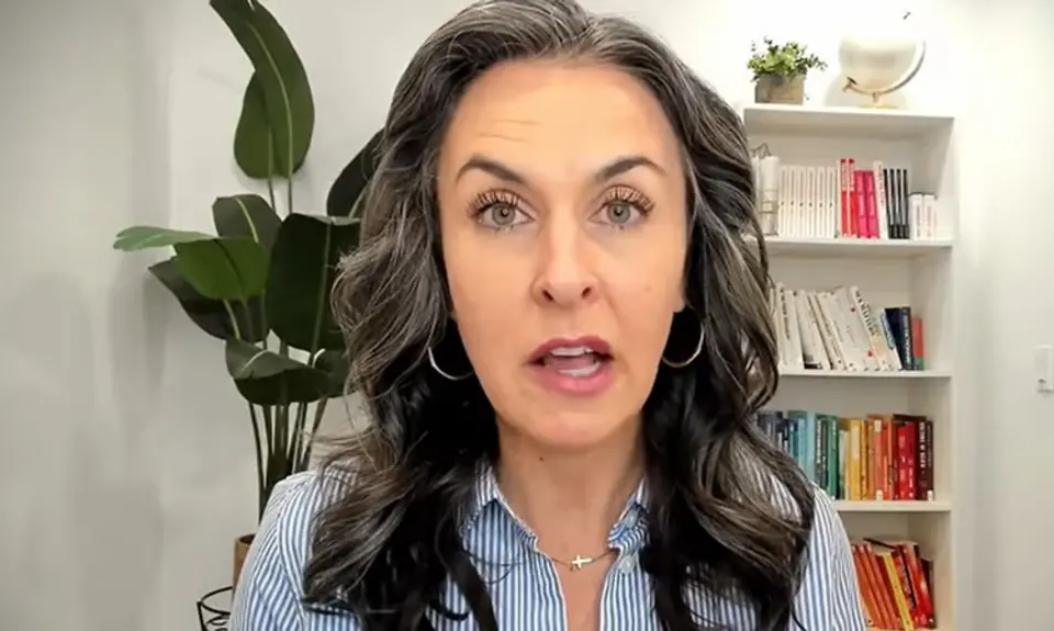 Katy Faust, a white woman with long black hair streaked with gray, speaks into the camera; a bookshelf and plant are visible behind her