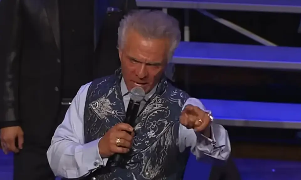 Kent Christmas, standing on stage at the Ryman Auditorium, speaks into a microphone in his right hand while pointing toward the crowd with his left hand; he looks angry.