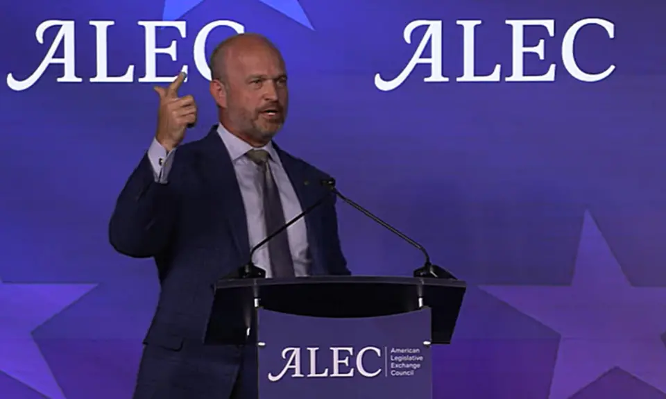 Heritage Foundation President Kevin Roberts, a bald white man wearing suit and tie, gestures with his right hand while speaking behind a lectern bearing the ALEC logo, which is also repeated on the background. 