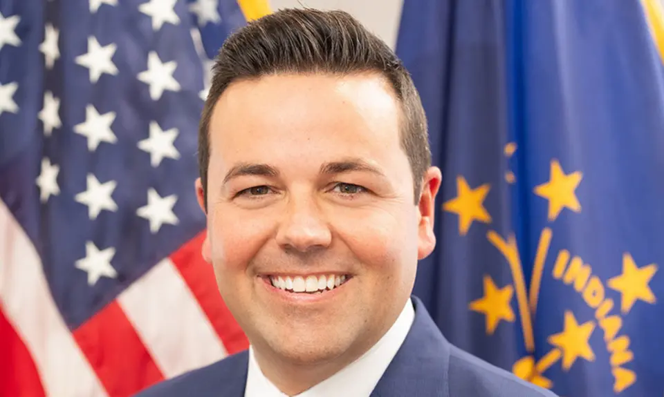 Portrait-style official photo of Indiana Lt. Gov. Micah Beckwith smiling into the camera with U.S. and Indiana flags visible behind him.