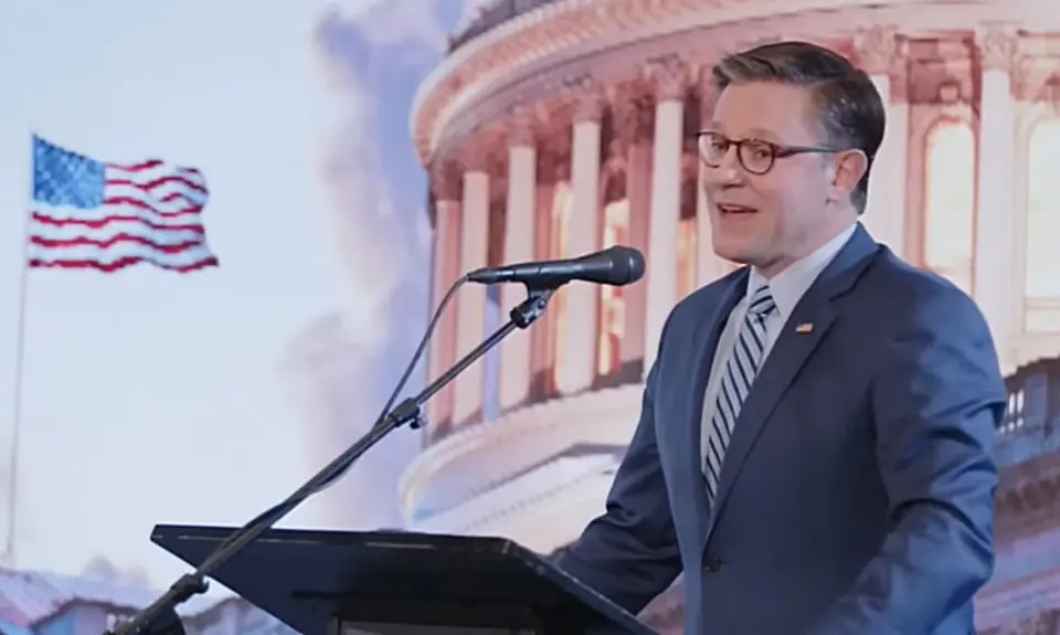 Image shows Mike Johnson standing and a lectern and speaking into a microphone in front of an illustrated image showing the Capitol dome and a U.S. flag fluttering in the breeze.