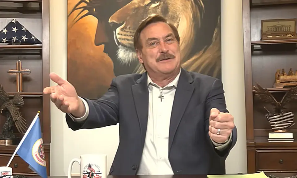 Mike Lindell, seated in front of a painting of a lion's head superimposed on the head of Christ, gestures with both hands. A cross and a US flag are visible on the shelves behind him. 