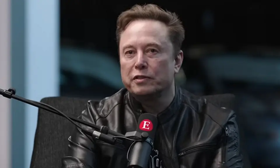 Elon Musk, in black shirt and leather jacket, is seated at a microphone that is emblazoned with a capital letter E, the logo for The Economist magazine