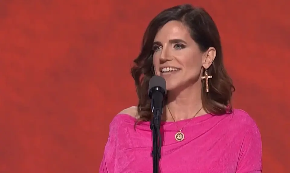 Rep. Nancy Mace, wearing bright pink dress and oversize cross-shaped earrings, speaks into a microphone in front of a red background on stage at the 2024 Republican National Convention.