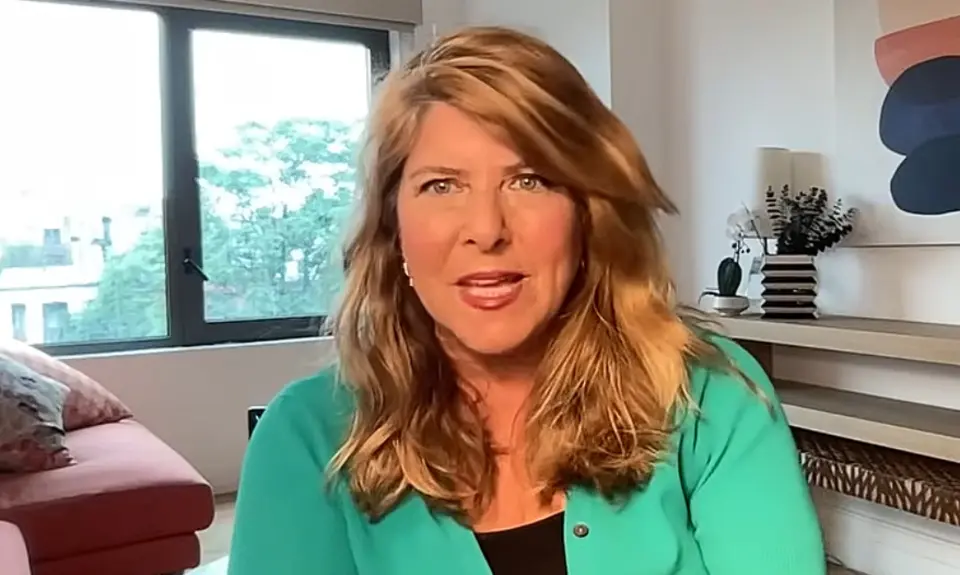 Naomi Wolf is seated in what appears to be a home office, with a window, shelf, and art visible behind her.
