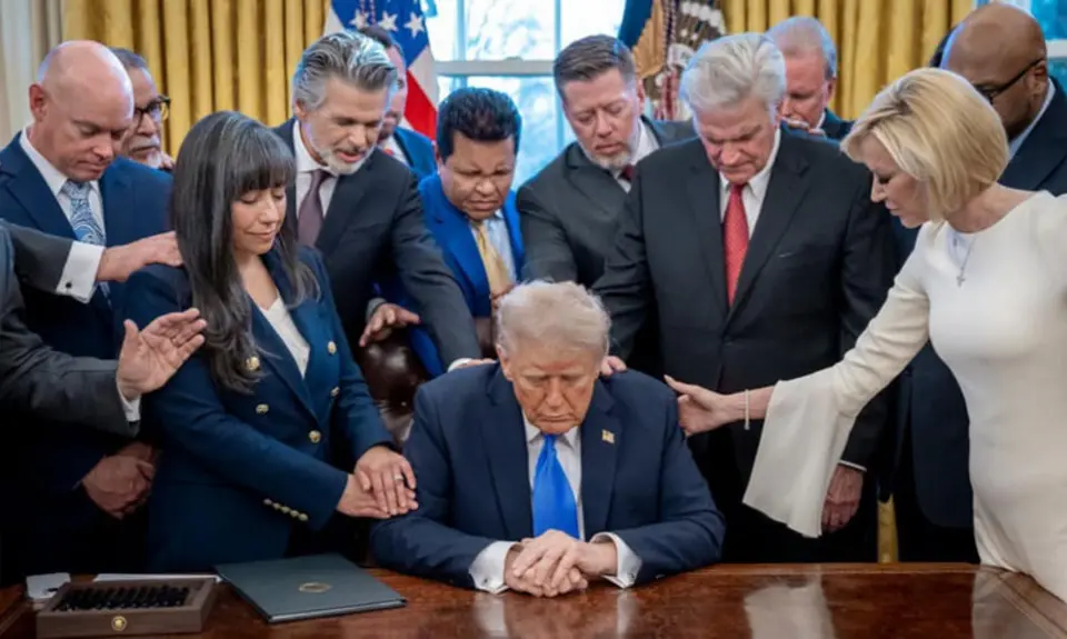 White House photo of Trump seated at his desk in the Oval Office surrounded by religious-right leaders; White House Faith Director Paula White, standing at right of image, lays her right hand on Trump's shoulder as she prays,