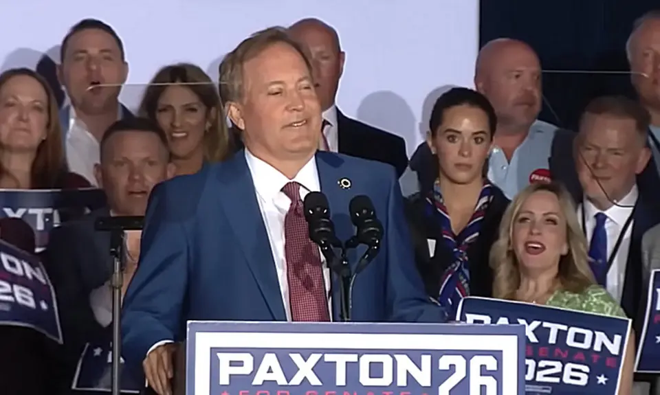 Texas Attorney General Ken Paxton stands at microphone behind a lectern with his Paxton 26 campaign logo. Supporters holding Paxton signs are gathered behind him.