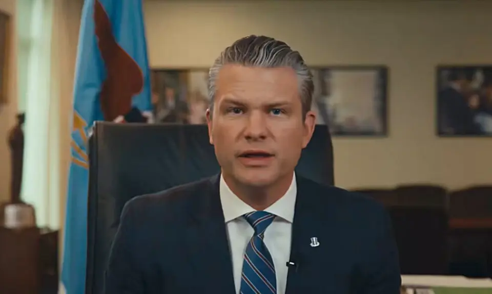 Pete Hegseth, wearing suit and tie, is seated in his office and speaking into the camera.