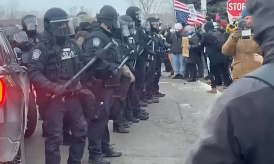 Image shows line of helmeted baton-wielding law enforcement officers facing off against protesters