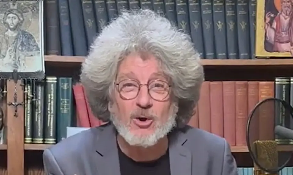 Randall Terry, white man with gray beard and full head of feathered gray hair, is seated at a microphone in front of bookshelves.