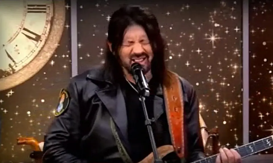Robin Bullock, a white man with long black hair, grimace while speaking/singing into a microphone and holding a guitar. 