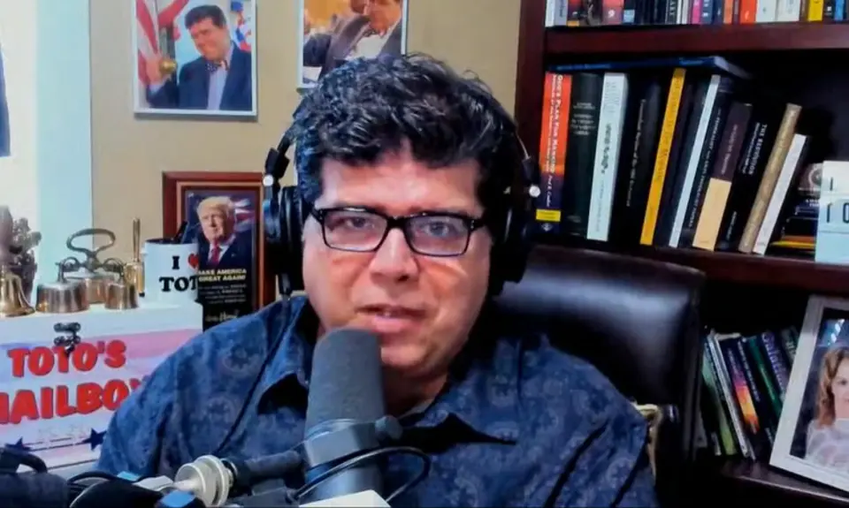 Shane Vaugh, wearing glasses and headphones, is seated in a cluttered office speaking into a microphone.