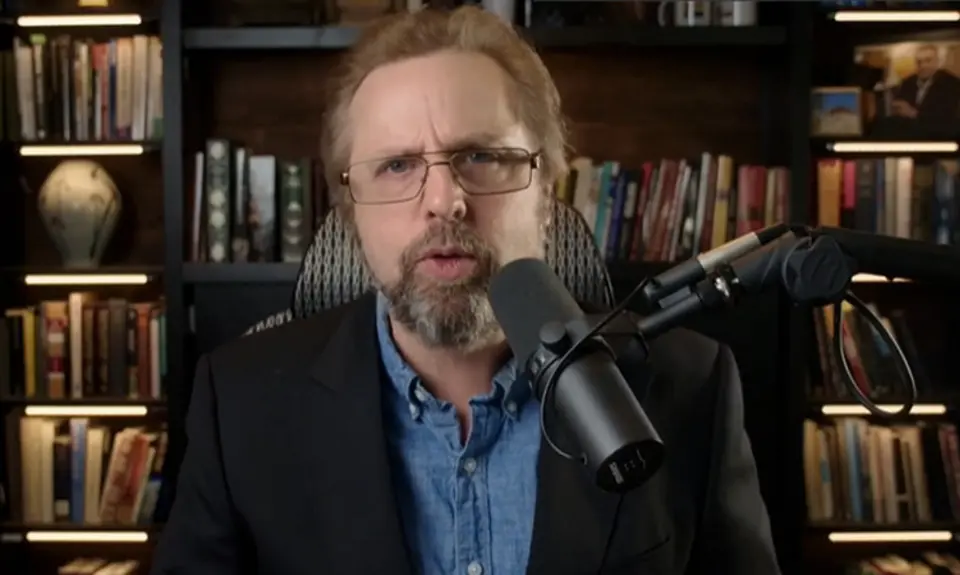 Steve Turley, a white man with red hair and beard, speaking into a microphone while seated in front of bookshelves in what appears to be a home office or library.