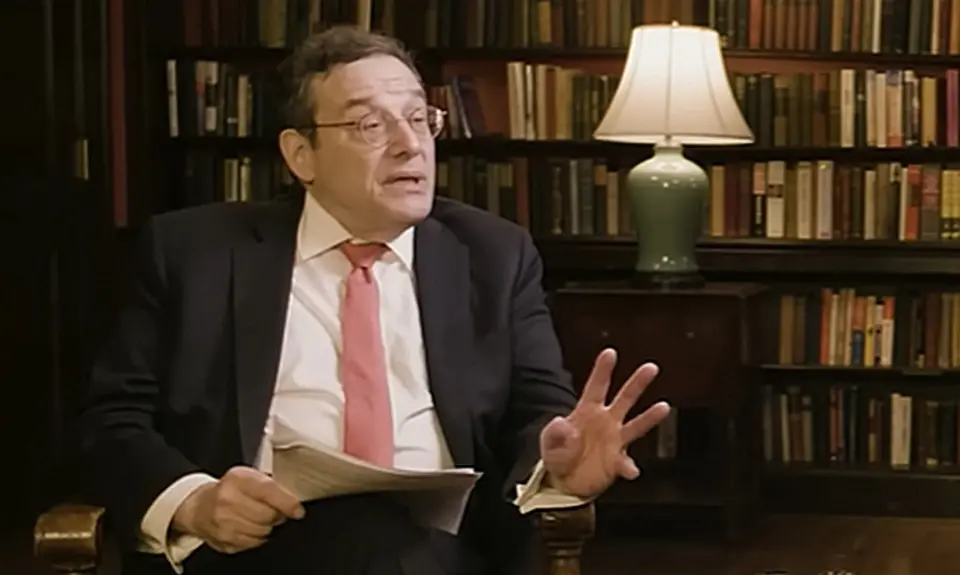 Tom Klingenstein, a white man wearing glasses and a suit and tie, holds papers in his right hand and gestures with his left. He is seated in front of bookshelves in what appears to be an office or home library. He is looking toward his left and speaking with John Eastman, who is not visible in this image cropped from video of their interview.