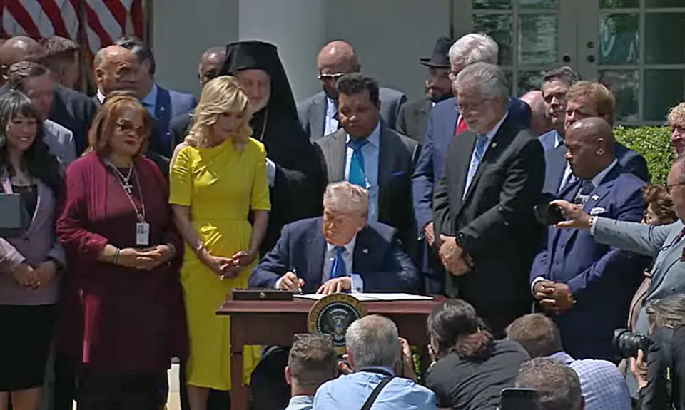 President Donald Trump seated at a desk in the White House rose garden, signing an executive order creating a Religious Liberty Commission, as White House aide Paula White and a group of religious leaders and activists look on. 