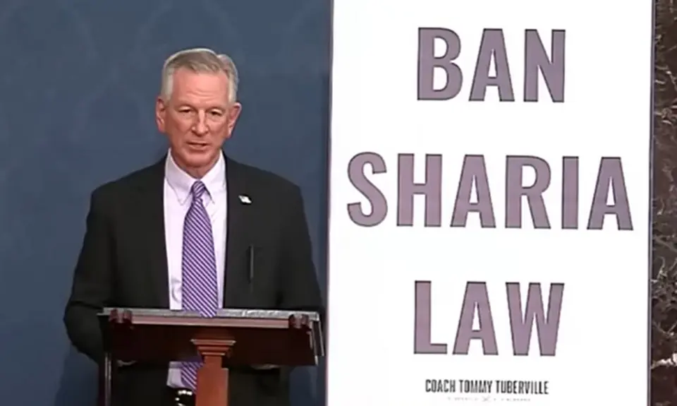 Sen. Tommy Tuberville, wearing suit and tie, stands behind lectern next to a huge poster reading BAN SHARIA LAW.