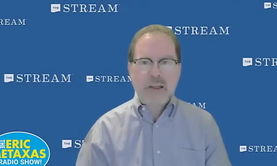 John Zmirak, a white man wearing a button-down shirt, is speaking in front of a background that repeats the logo for religious-right media outlet The Stream. The logo for The Eric Metaxas radio show appears in the lower left-hand corner of the screen. 