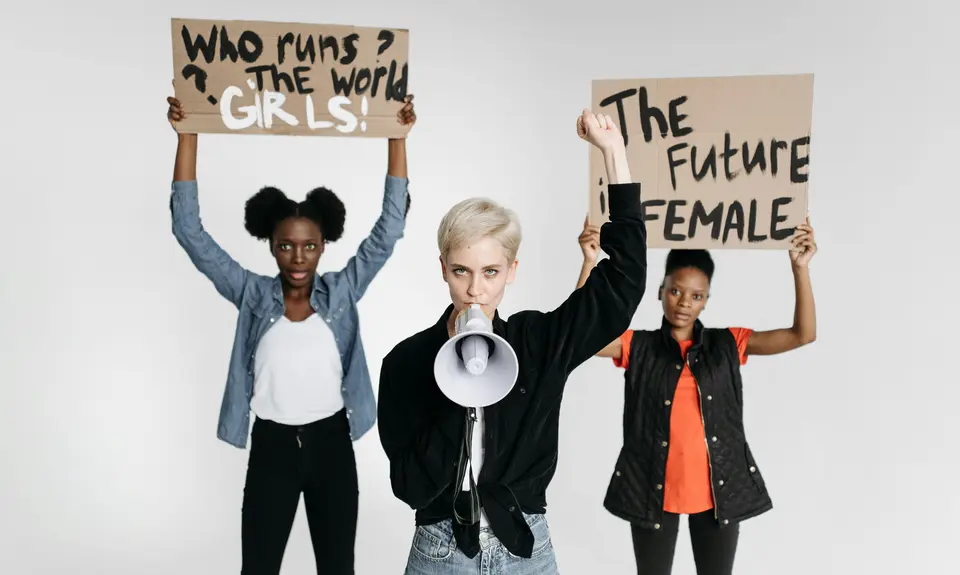 Three women, one holding a sign reading "who runs the world? Girls!", the second woman holding a megaphone, and the third holding a sign reading "the future is female."