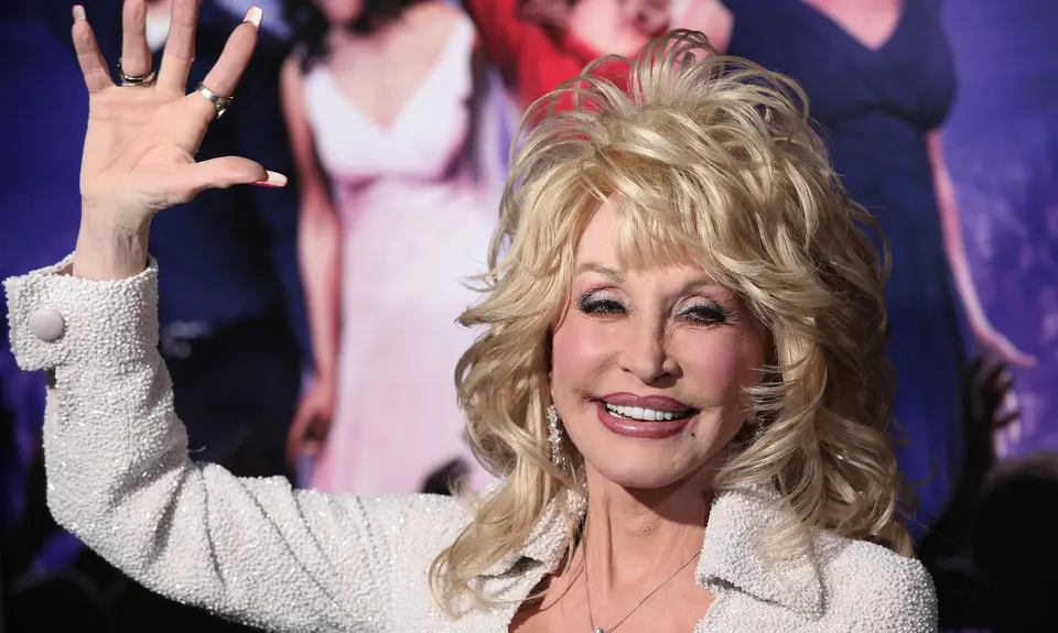 Dolly Parton, a white woman with blonde hair, wearing a white jacket and smiling and waving.