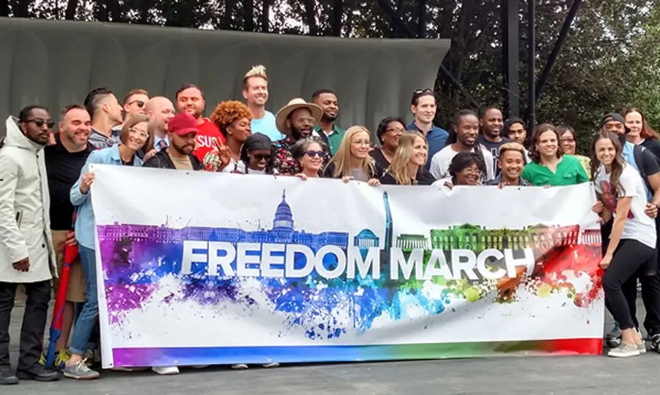 A couple dozen speakers and singers gather on stage behind a rainbow-colored "Freedom March" banner.