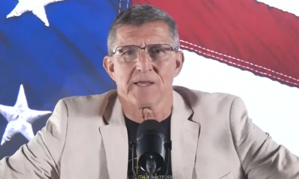 Michael Flynn speaks into a microphone seated in front of a close-up image of stars and stripes from a U.S. flag. 