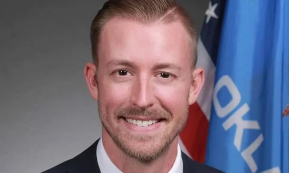 Official-style portrait of Ryan Walters smiling into the camera with US and Oklahoma flags visible over his left shoulder.
