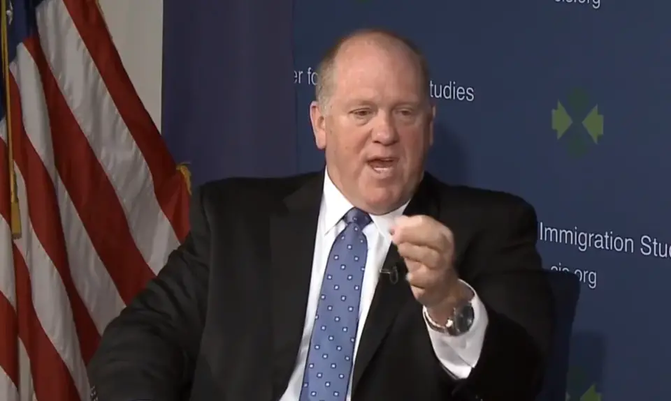 Tom Homan, in suit and tie and seated in front of an American flag, gestures while speaking. 